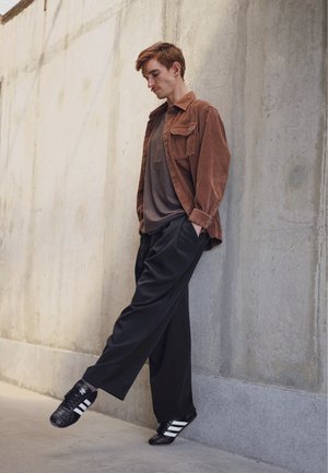 Camisa de pana marrón, camiseta gris debajo, pantalones anchos negros y zapatillas negras con rayas blancas. El fondo presenta una pared de concreto texturizado.