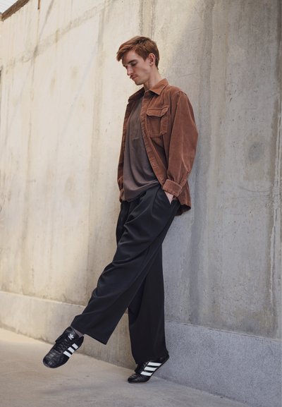 Camisa de pana marrón, camiseta gris debajo, pantalones anchos negros y zapatillas negras con rayas blancas. El fondo presenta una pared de concreto texturizado.