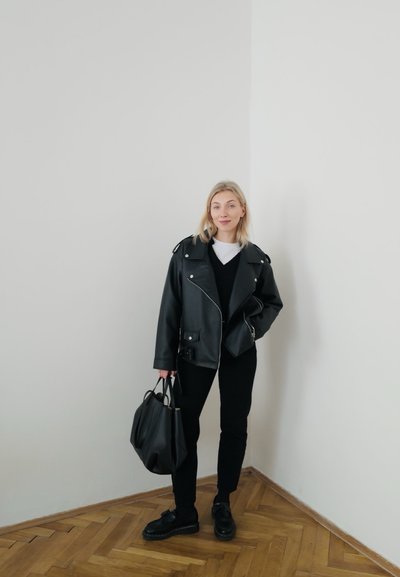 Chaqueta de cuero negro de motorista sobre un suéter negro, pantalones negros y zapatos negros. Sosteniendo un bolso negro. Suelo de madera clara y pared lisa.