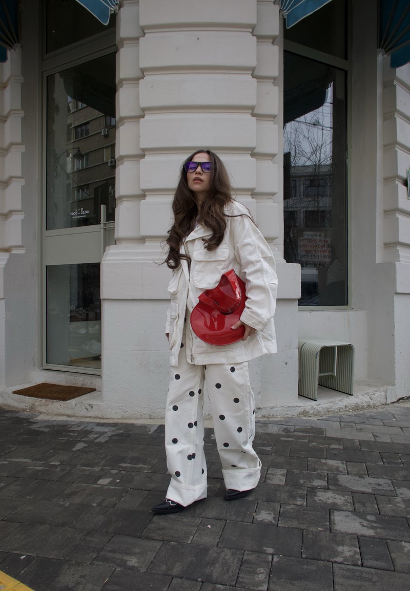 Donna con giacca bianca e pantaloni a pois che tiene una borsa rossa, in piedi su una strada acciottolata davanti a un edificio bianco con grandi finestre.