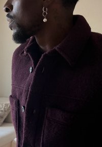 Homme portant un manteau en laine bordeaux foncé avec des boutons noirs et une boucle d'oreille argentée en forme d'anneau avec une perle pendante à l'oreille gauche.