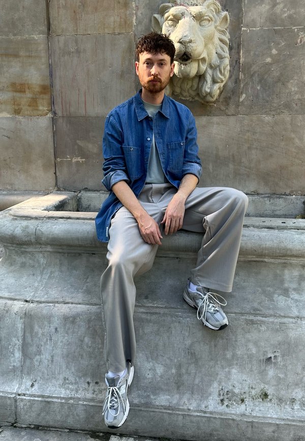 Homme aux cheveux bouclés foncés et barbe assis sur un banc en pierre sous une sculpture de tête de lion, portant une veste en jean, un pantalon clair et des baskets.