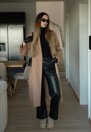 Mujer con cabello largo que lleva gafas de sol negras, suéter de cuello alto negro, pantalones de cuero, abrigo beige, guantes y botas, parada en un pasillo de cocina moderno.