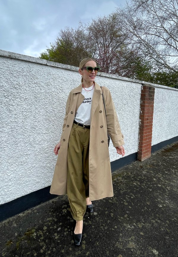 Femme portant un trench beige, un pantalon vert olive, une chemise blanche avec du texte, des talons noirs et des lunettes de soleil qui marche sur un trottoir près d'un mur blanc.