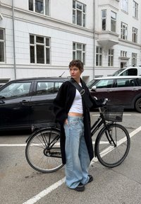 Czarna rower z metalowym koszem i szerokimi oponami. Osoba ubrana w długi czarny płaszcz, biały krótki top i jasnoniebieskie szerokie spodnie.