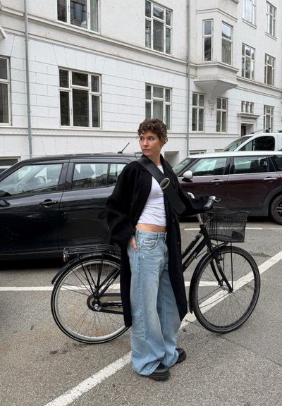 Bicicleta negra con una cesta de metal y neumáticos anchos. Persona vestida con un abrigo negro largo, top corto blanco y jeans de pierna ancha azul claro.