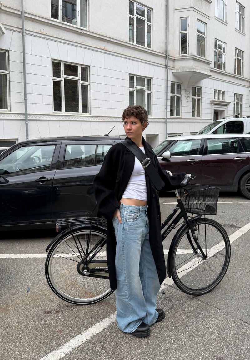 Bicicletta nera con un cestino metallico e pneumatici larghi. Persona che indossa un lungo cappotto nero, una maglietta bianca corta e jeans larghi blu chiaro.