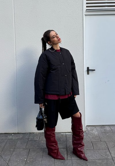 Mujer con el cabello trenzado, llevando una chaqueta negra acolchada, pantalones cortos negros, grandes pendientes de aro, botas burgundy hasta la rodilla y sosteniendo un bolso negro.