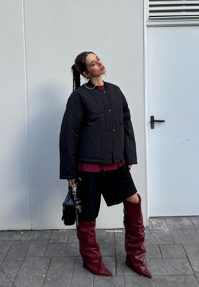 Femme aux cheveux tressés portant une veste matelassée noire, un short noir, de grandes boucles d'oreilles, des bottes bordeaux montantes jusqu'aux genoux, tenant un sac à main noir.