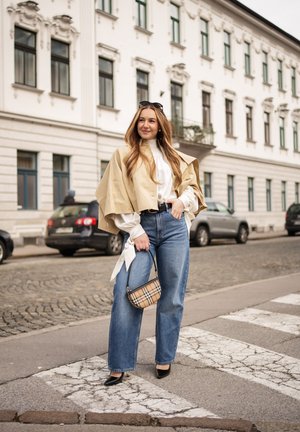 Mujer de pie en un paso de peatones de empedrado, vestida con capa beige, blusa blanca, vaqueros azules, tacones negros, gafas de sol sobre la cabeza y llevando un bolso de cuadros.