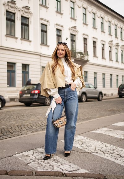Mujer de pie en un paso de peatones de empedrado, vestida con capa beige, blusa blanca, vaqueros azules, tacones negros, gafas de sol sobre la cabeza y llevando un bolso de cuadros.