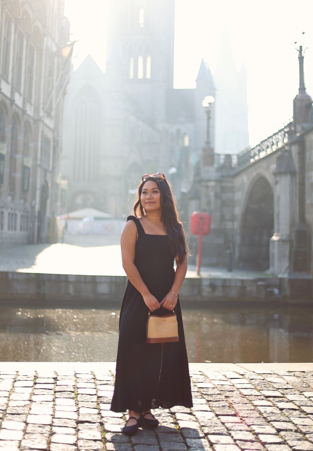 Sort maxikjole med firkantet udskæring, parret med en vævet håndtaske. Blødt sollys belyser scenen med en brostensbelagt gade som baggrund.