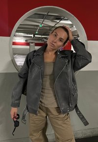 Γκρι δερμάτινο μπουφάν με φθαρμένη υφή, φαρδιά εφαρμογή και έντονη ραφή. Συνδυασμένο με μπεζ μπλούζα και χαλαρές καμηλό παντελόνες.