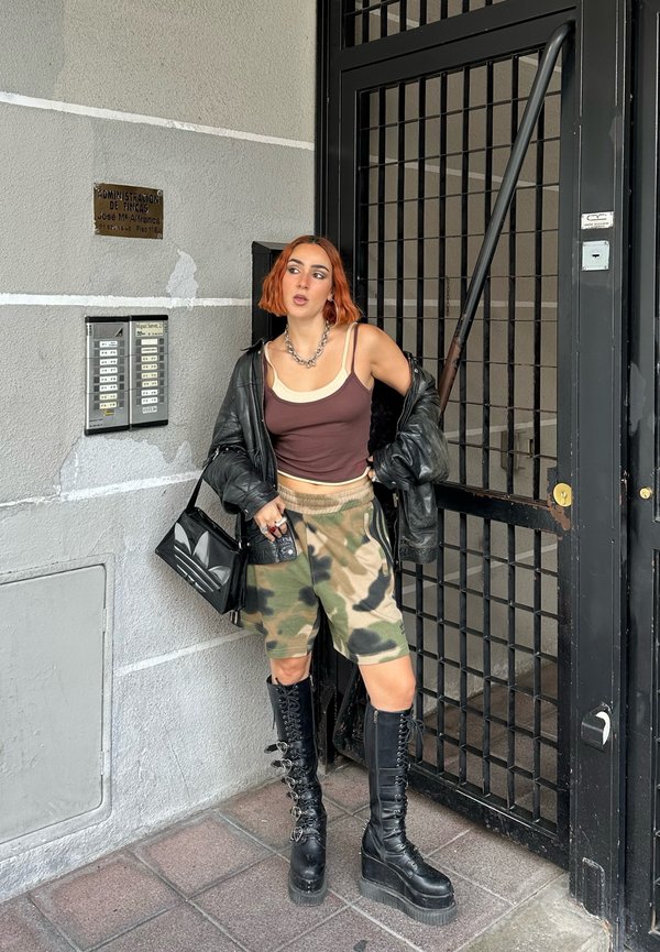 Chaqueta de cuero negra, top de tirantes marrón, pantalones cortos de camuflaje, botas negras hasta la rodilla con cordones y bolso negro. El fondo presenta una puerta de metal.