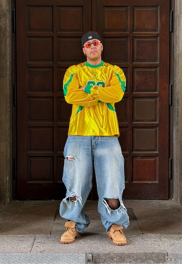 Uomo con cappellino nero, occhiali da sole arancioni, maglia sportiva gialla, jeans larghi strappati e stivali beige, in piedi con le braccia incrociate davanti a una porta in legno con pannelli.