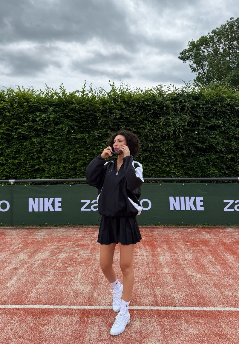 Femme en tenue de sport noire ajustant ses lunettes de soleil sur un court de tennis en terre battue avec des banderoles Nike et Zalando derrière une haie verte.
