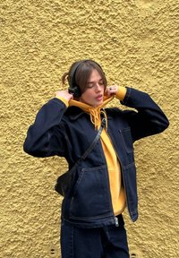 Jeansjack over een gele hoodie, zwarte headphones en een kleine crossbody tas. Geplaatst tegen een getextureerde gele muur.