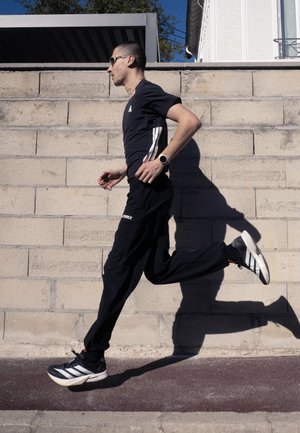 Hombre con gafas de sol y ropa deportiva negra corriendo junto a una pared de hormigón en un día soleado.