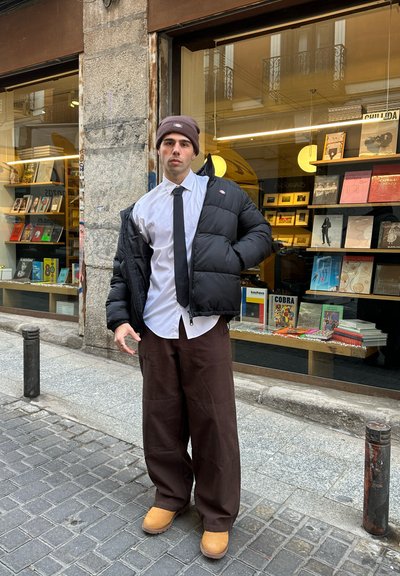 Chaqueta de abrigo negra, camisa blanca, corbata negra, pantalones anchos marrones y botas color miel. De pie frente a una librería con estanterías llenas de libros.