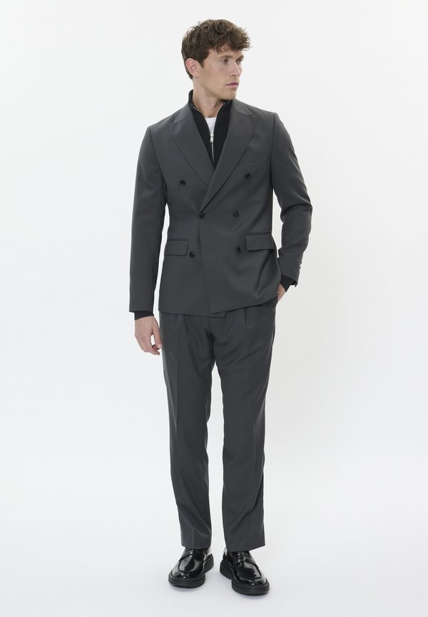 Completo grigio doppiopetto con vestibilità sartoriale, dotato di rever a punta e tasche laterali, abbinato a scarpe nere lucide.
