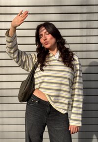 Mladá žena s dlhými tmavými vlasmi v pásikovanom tričku a čiernych džínsoch, zakrývajúca si rukou oči, stojaca pred kovovou roletou.