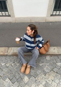 Ein blauer und grauer gestreifter Cardigan, graue Schlagjeans, hellbraune Ankle Boots, hält eine Kaffeetasse, mit einer braunen Tasche daneben auf Kopfsteinpflaster.