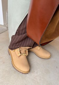 Tan veterschoenen met gouden oogjes en gestructureerde zolen, gecombineerd met bruine gestreepte broek en een groene top. Een bruine leren tas is zichtbaar.