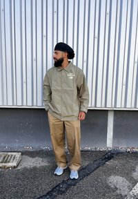 Ελαιόχρωμο αδιάβροχο μπουφάν με κουμπιά και λογότυπο, μπεζ παντελόνι με φαρδιά πόδια, γκρι αθλητικά παπούτσια, στέκεται μπροστά σε τοίχο από λωρίδες μετάλλου.