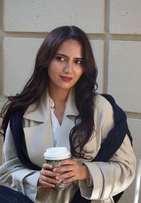 Beiger Trenchcoat mit breiten Revers, darüber ein marineblauer Strickschal in Lagen. Hält eine Kaffeetasse mit einem gemusterten Tassenbecher. Lange braune Haare.