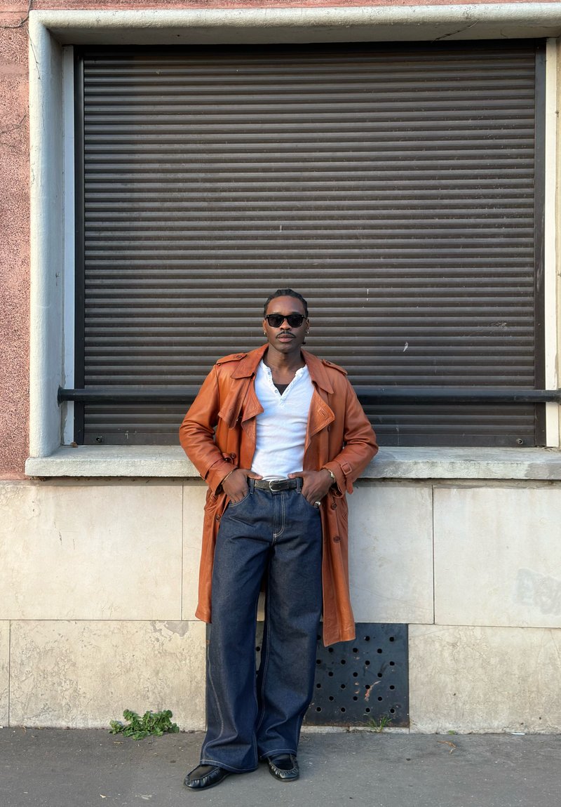 Homem usando óculos de sol, camisa branca, casaco trench coat de couro castanho e jeans de pernas largas, com as mãos nos bolsos, encostado numa porta de loja fechada.