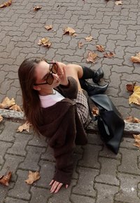 Smeđi, pretjerano veliki pleteni džemper, bijela majica, karirana suknja, crne čizme do koljena, velika crna torba i tamne sunčane naočale s ravnim staklom.