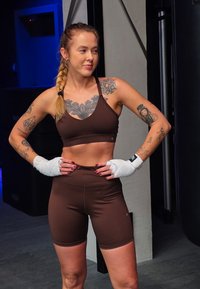 Haut sportif court marron et short assorti, texture du tissu visible. Les mains sont enveloppées de gaze blanche, décorées de divers tatouages.