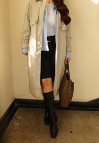 Beige trench kabát, fehér bodysuit, csíkos kék ing, fekete sort, térdig érő fekete csizmák és egy szegecses barna táska.