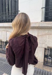 Blusa color bordeaux scuro con maniche lunghe, tessuto testurizzato, fiocco annodato sul retro e vestibilità rilassata, abbinata a pantaloni bianchi.