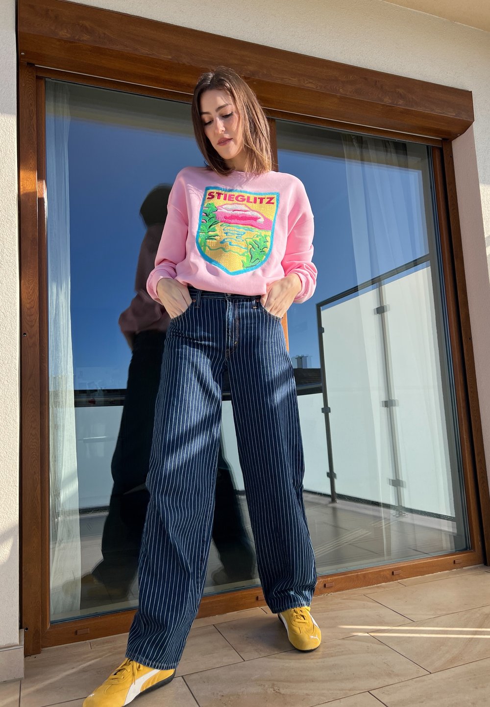 Sweatshirt cor-de-rosa com o texto "STIEGLITZ" e um design gráfico; jeans largas listradas em azul-marinho e ténis amarelos. De pé numa varanda.