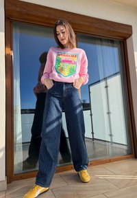 Sweatshirt cor-de-rosa com o texto "STIEGLITZ" e um design gráfico; jeans largas listradas em azul-marinho e ténis amarelos. De pé numa varanda.