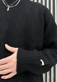 Personne portant un pull en maille noire avec un collier chaîne et un pendentif en plume, main posée sur la poitrine, petite étiquette en tissu sur le poignet de la manche.