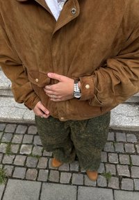 Brązowa zamszowa kurtka z zatrzaskami, noszona na białej koszuli; w połączeniu z zielonymi spodniami w wzory i brązowymi butami. Srebrny zegarek z zapięciem.