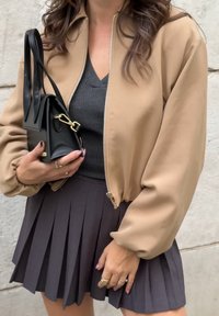 Una chaqueta bomber beige con cremallera, llevada sobre un suéter de pico gris oscuro y una minifalda negra con pliegues, sosteniendo un pequeño bolso negro.