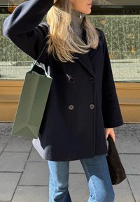 Dvigubos užsegimo mėlynas paltas su dideliais mygtukais, derinamas su pilka aukštu kaklu. Laikomas tamsiai žalias popierinis maišelis ir juoda tekstūruota rankinė.