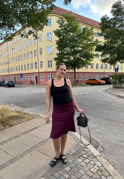 Camiseta negra sin mangas, falda burdeos hasta la rodilla, zapatos negros. Sosteniendo un pequeño bolso marrón. Escena callejera con edificios y coches al fondo.