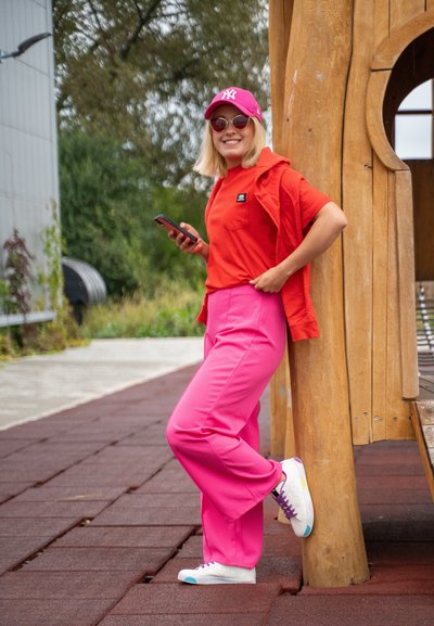 Camiseta roja de gran tamaño, pantalones rosas de pierna ancha, zapatillas blancas con acentos coloridos y gorra rosa con un logo, de pie junto a una estructura de madera.
