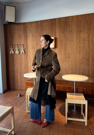 Mujer con abrigo marrón, falda negra de capas, vaqueros azules y zapatos rojos sostiene un pastel en una cafetería de madera con mesas redondas y taburetes.