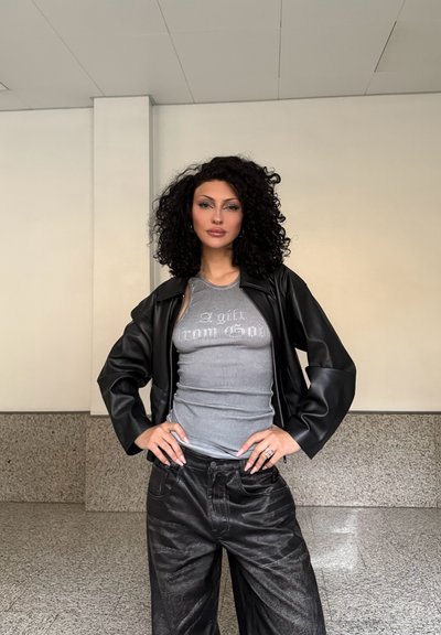 Chaqueta de cuero negra sobre una camiseta de tirantes gris con relieve, combinada con pantalones holgados negros. El cabello es rizado; el entorno presenta un fondo minimalista.