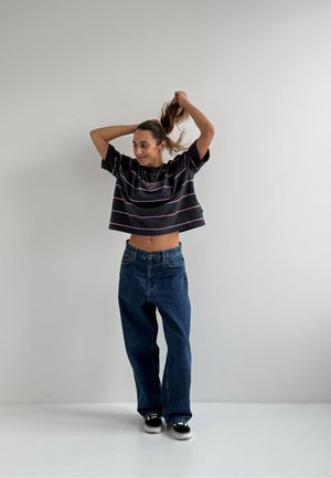 Camiseta negra recortada con rayas rosas, jeans de mezclilla azul holgados y zapatillas negras; modelo juega con su cabello frente a un fondo liso.