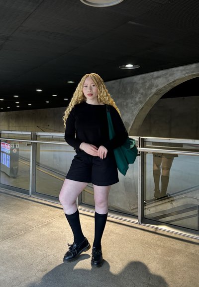 Top negro de manga larga, pantalones cortos negros, calcetas hasta la rodilla y zapatos negros. Sostiene una bolsa de mano verde. Fondo de concreto con superficies reflectantes.