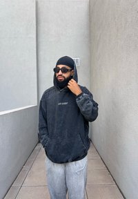 Μαύρη oversized φούτερ με τσέπη μπροστά και λογότυπο, συνδυασμένη με ανοιχτού γκρι παντελόνι. Λείο ύφασμα με φθαρμένη όψη. Φοράει γυαλιά ηλίου.