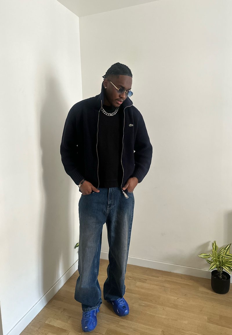 Homme portant une veste noire, une chemise noire, un jean bleu et des baskets bleues, se tenant à l'intérieur près d'une plante sur un sol en bois clair.