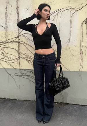 Mujer de cabello corto y negro, que lleva un top negro corto y jeans oscuros, sosteniendo un bolso negro, de pie frente a una pared clara con enredaderas trepadoras.