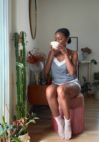 Graues Tanktop mit weißer Spitzenborte, kombiniert mit hellgrauen Shorts. Sitzt auf einem pinkfarbenen, fransigen Hocker und hält eine cremefarbene Tasse.